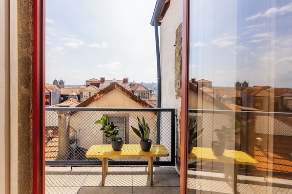 Ein Balkon mit einem gelben Tisch und zwei Blumentöpfen, mit Blick auf eine Stadtlandschaft mit traditionellen Dächern.