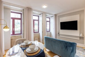 Ein Essbereich mit einem Tisch für vier Personen, einem blauen Sofa, einem an der Wand befestigten Fernseher und großen Fenstern mit Vorhängen.