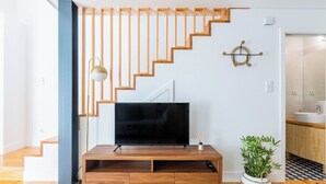 Un salon moderne avec un escalier en bois, un téléviseur à écran plat posé sur un meuble en bois, une plante en pot et une décoration murale minimaliste.