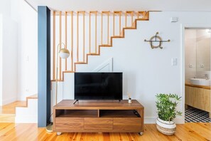 Ein modernes Wohnzimmer mit einer Holztreppe, einem Flachbildfernseher auf einem Holzboden, einer Topfpflanze und schlichter Wandgestaltung.