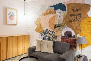 Ein gemütliches Wohnzimmer mit grauem Sofa, einem Holzschrank und einer Wandmalerei mit dem Schild 'Our happy place'.
