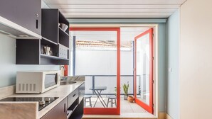 Een kleine keuken met een magnetron, spoelbak en kastjes, die uitkomt op een balkon met een tafel en stoelen.