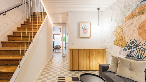 Een trap met houten treden en een witte leuning, die leidt naar een kamer met een zwart-witte vloer, een bank en een kleine tafel.