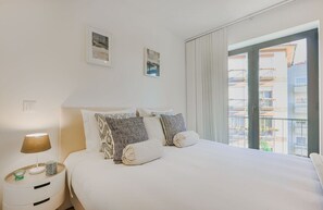 Ein Hotelzimmer mit einem Bett, einem Nachttisch mit Lampe, zwei gerahmten Bildern an der Wand und Blick auf Gebäude durch das Fenster.