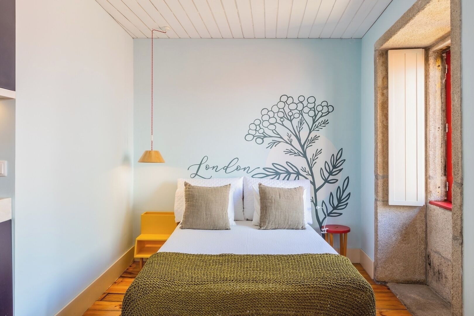 A bedroom with a bed, pillows, a hanging lamp, a wall mural, and a wooden floor.