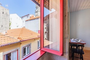 Ein Zimmer mit großem Fenster und Blick auf die Stadt, ein kleiner Tisch mit Stuhl sowie eine Ansicht der Nachbargebäude.