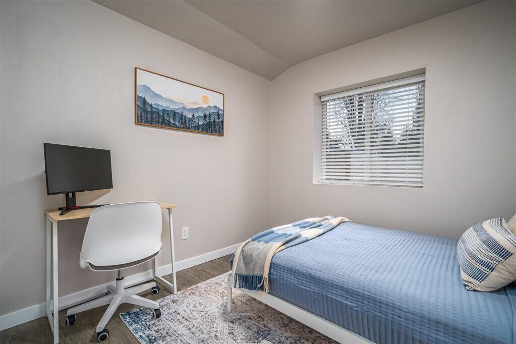 A bedroom with a bed, a desk with a computer, a chair, a window with blinds, and a painting on the wall.