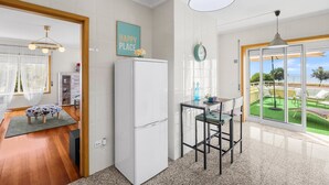 A modern kitchen with a white refrigerator, a dining area with a table and chairs, and a view of a green lawn through a glass door. - São Félix Ocean Views | 1BR & Terrace (São Félix da Marinha)