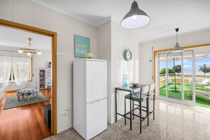 Cozinha moderna com geladeira branca, área de refeições com mesa e cadeiras, e vista para um gramado verde através de uma porta de vidro.