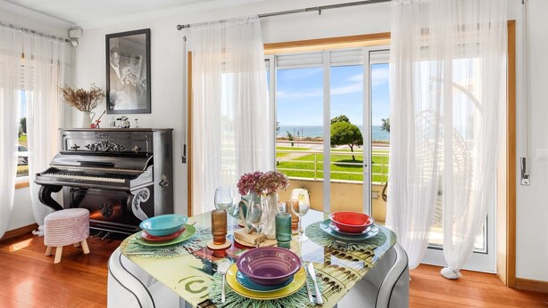 A dining area with a piano, a table set with colorful plates and glasses, and a view of a coastal landscape through large glass doors. - São Félix Ocean Views | 1BR & Terrace (São Félix da Marinha)