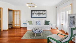 A modern living room with a wooden floor, a white sofa, a green armchair, a patterned ottoman, and a ceiling light fixture. - São Félix Ocean Views | 1BR & Terrace (São Félix da Marinha)