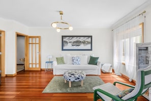 Uma sala de estar moderna com piso de madeira, um sofá branco, uma poltrona verde, um banquinho estampado e um lustre.