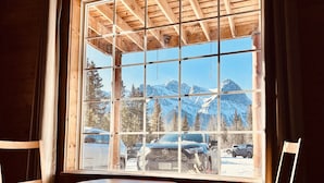 Una habitación con un ventanal amplio que ofrece vistas a montañas nevadas y coches aparcados. Hay una mesa redonda de madera y dos sillas de madera con cojines rojos.