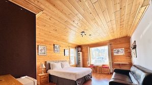 Una habitación acogedora con paredes y techo de madera, una cama con cabecera gris, un sofá de cuero negro, una mesita y una ventana con vistas a árboles.