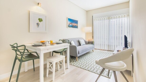 Ein modernes Wohnzimmer mit einer Couch, einem Esstisch mit Stühlen, einer Lampe und Blick durch ein Fenster.