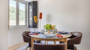 A dining area with a round table, four chairs, a vase with flowers, and a window with blinds. - Rosarinho Cruz invites to her Atelier by the Sea (Porto)