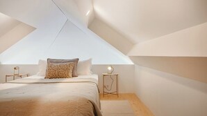 A bedroom with a sloped ceiling, a bed with a beige bedspread, two bedside tables with lamps, and a small rug on the floor. - Rosarinho Cruz invites to her Atelier by the Sea (Porto)