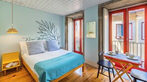 Ein Schlafzimmer mit einem Bett, einem Schreibtisch mit Stühlen, einer Hängeleuchte und Blick ins Freie.