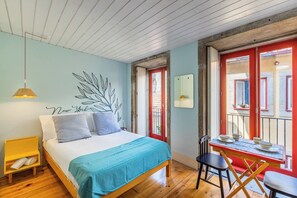 Ein Schlafzimmer mit einem Bett, einem Schreibtisch mit Stühlen, einer Hängeleuchte und Blick ins Freie.