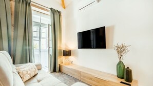 Ein Wohnzimmer mit einer Couch, einem flachbildfernseher an der Wand, einem Holztischelement und einer Vase mit getrockneten Blumen.