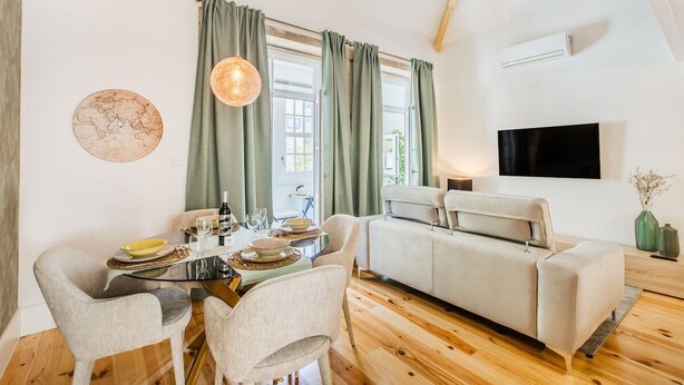 Ein modernes Wohnzimmer mit Sofa, Esstisch, Stühlen, einer Hängeleuchte und einem wandmontierten Flachbildfernseher.