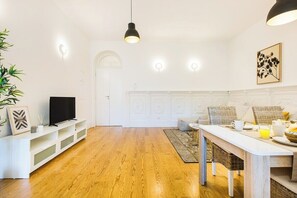 Ein modernes Wohnzimmer mit Holzboden, weißen Möbeln, einem Fernseher und einem Essbereich mit Tisch für zwei Personen.