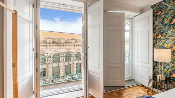 A room with a wooden floor, a bed, a window with a view of a building, and a door leading to another room. - São Bento Deluxe 42 with Balcony (Porto)