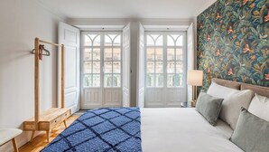 A bedroom with a bed, a bench, a wooden hanger rack, a window with a view, and a patterned wall. - São Bento Deluxe 42 with Balcony (Porto)