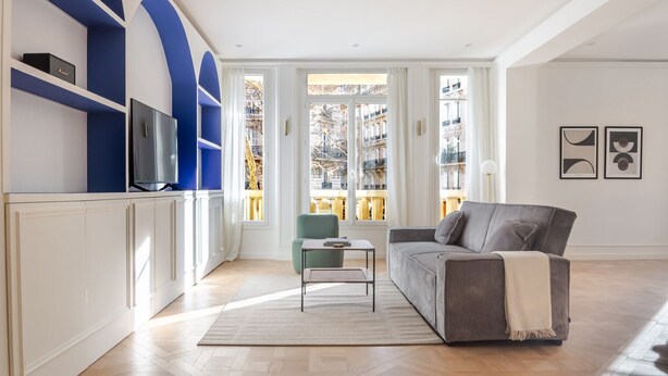 Una sala moderna con un sofá, una mesita y un televisor sobre un mueble blanco. Amplias ventanas ofrecen vistas a edificios.