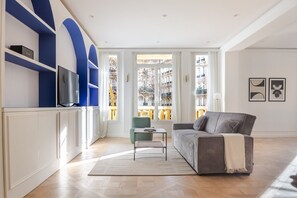 Ein modernes Wohnzimmer mit einer Couch, einem kleinen Tisch und einem Fernseher auf einem weißen Schrank. Große Fenster bieten einen Blick auf die Gebäude gegenüber.