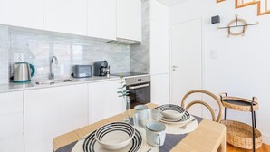 Ett kompakt kök med vita skåp, en väggbeklädnad av marmor, en inbyggd ugn och en matplats med ett bord och stolar i trä.