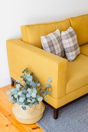 Ein senfgelbes Sofa mit karierten Kissen, ein gewebter Korb mit Eukalyptusblättern und ein grauer Teppich.
