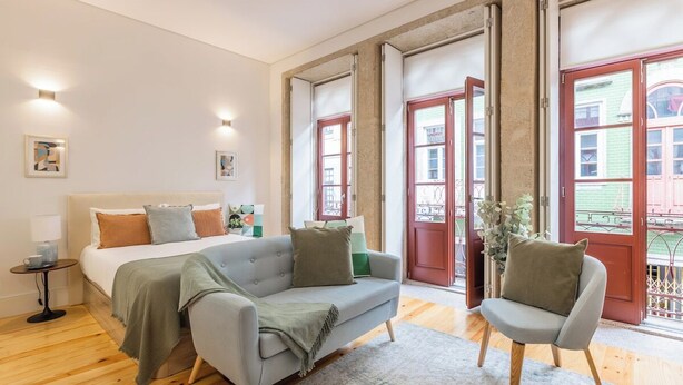 Une chambre à coucher comprenant un lit, un canapé, une chaise, une table de chevet et une fenêtre donnant sur des immeubles.