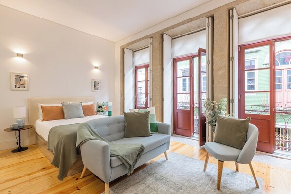 A bedroom with a bed, a sofa, a chair, a bedside table, and a window with a view of buildings. - Sol Downtown Studio with Juliet Balcony (Porto)
