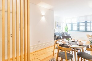 A dining area with a wooden table set for four, a sofa, and a window with blinds. - Bonfim Perfect Stays (Porto)