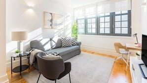 Una sala moderna con un sofá gris, una mesita redonda con una lámpara, un mueble para televisión y un ventanal con vistas.