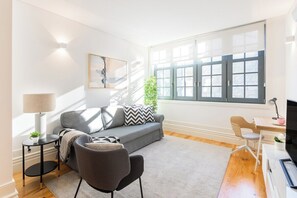 A modern living room with a grey sofa, a small round table with a lamp, a TV stand, and a window with a view. - Bonfim Perfect Stays (Porto)