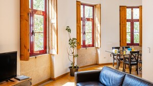 A room with a TV, a dining table with chairs, a potted plant, and large windows with wooden frames. - Casa Fernando @ Trindade (Porto)