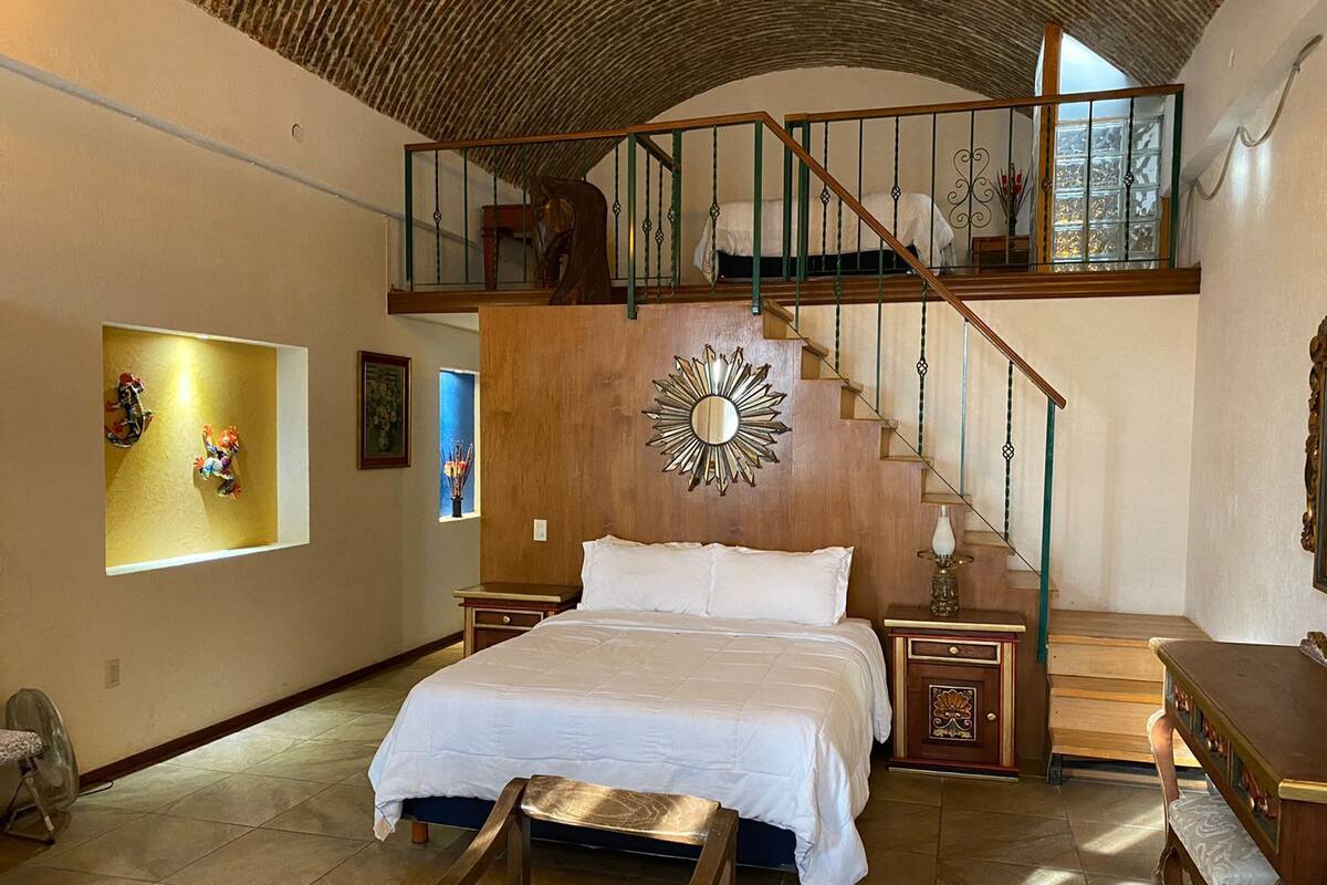 A bedroom with a large bed, a wooden headboard, a staircase, and a view of the outside.