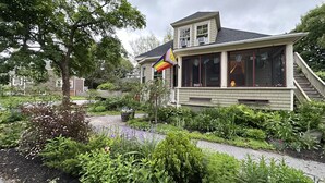 Une maison avec un porche abrité, un jardin avec différentes plantes et un drapeau arc-en-ciel.