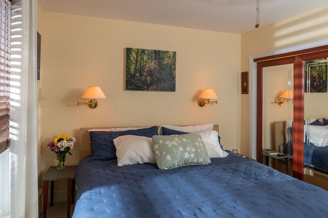 A bedroom with a large bed, two wall-mounted lamps, a painting, and a mirror.