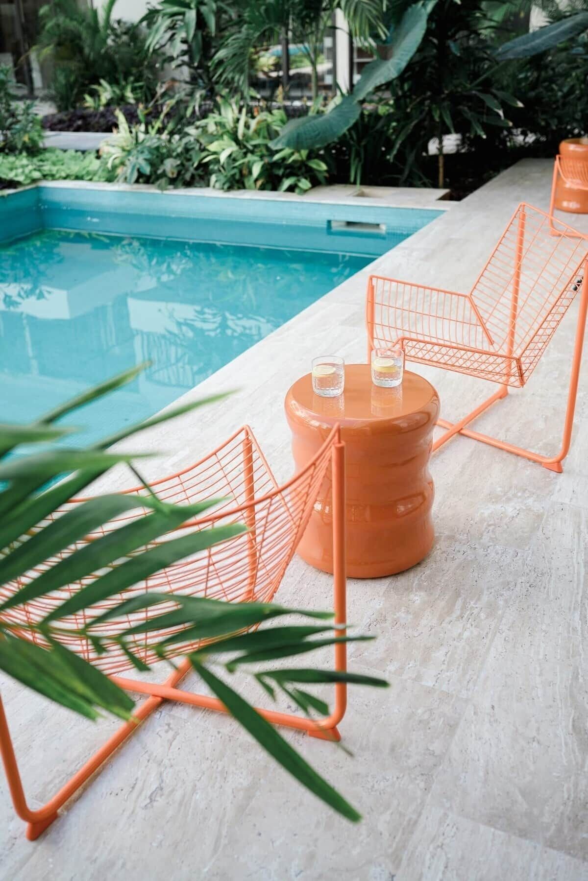 Ein Poolbereich mit weiß gefliestem Deck, einem runden, orangefarbenen Pflanzgefäß mit einem Glas und zwei orangefarbenen Drahtstühlen.