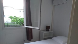 Ein kleines, sauberes Zimmer mit einem Einzelbett, einem Nachttisch mit Lampe und einem mit Vorhängen versehenen Fenster.
