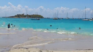 Ein Strand mit klarem türkisblauem Wasser, Booten und einer kleinen Insel im Hintergrund.