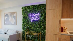 Una sala de estar moderna con un letrero de neón que dice "Miami Pulse", una pared verde con follaje, un sofá blanco, una mesa de centro dorada y un mueble de madera.