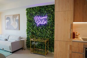 Ein modernes Wohnzimmer mit einem Neonlichtschild mit der Aufschrift 'Miami Pulse', einer grünen Wand mit Pflanzen, einem weißen Sofa, einem goldenen Couchtisch und einem Holzschrank.