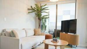 A modern living room with a sofa, a coffee table, a television, and a large window. - 2BR/2BA Palm Oasis at The Crosby (Miami)