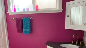 Un bany amb una paret de color rosa, mobles blancs, una finestra amb ampolles i un vàter.