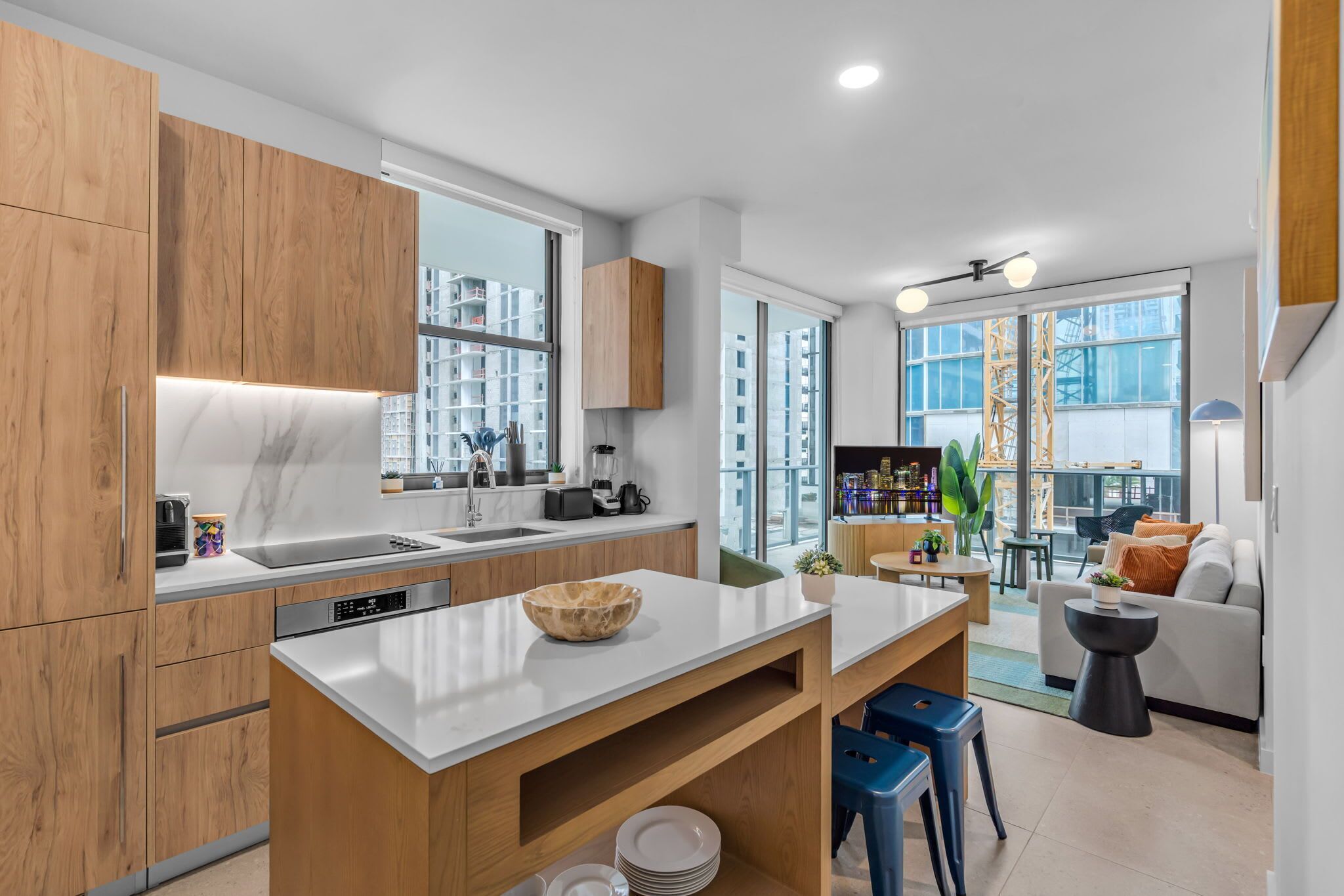 A modern kitchen with wooden cabinets, a white countertop, and a built-in dishwasher. There is a dining area with a table and chairs, and a sofa in the background.
