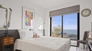 Een slaapkamer met een groot bed, een nachtkastje, een stoel, een raam met uitzicht op zee, en een klok aan de muur.